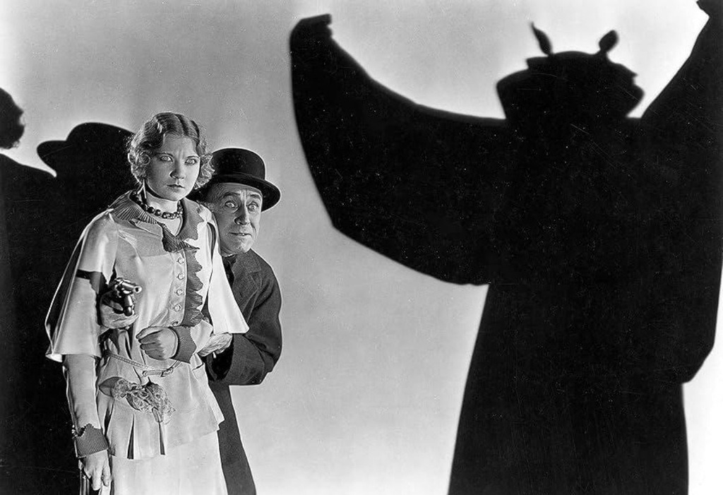 Extrait du film The Bat Whisper avec une femme, devant laquelle se trouve un homme qui tient un pistolet, et font face à une silhouette noire qui surgit devant eux.