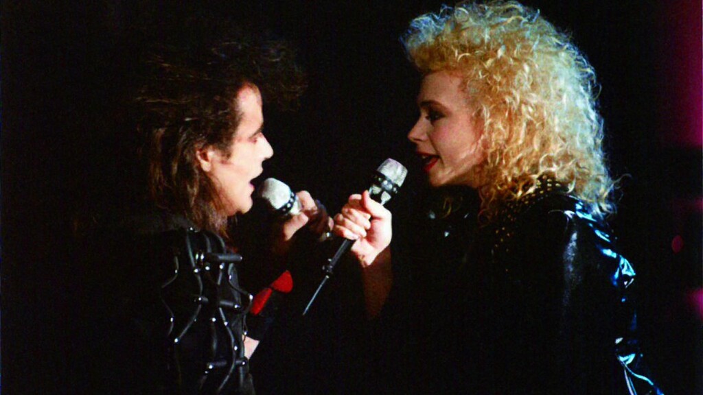 Un homme et une femme, tous deux en blousons de cuir noirs et avec des cheveux longs et frisés typiques des années 80, qui chantent ensemble.