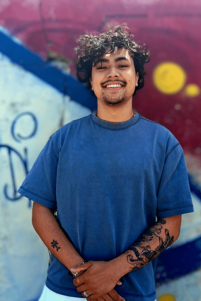 Arthur Khong, jeune homme aux cheveux bouclés et aux yeux noirs, de larges sourires et une moustache. Il porte un tee-shirt bleu et est adossé à un mur sur lequel il y a des graffitis moches, mais colorés. 