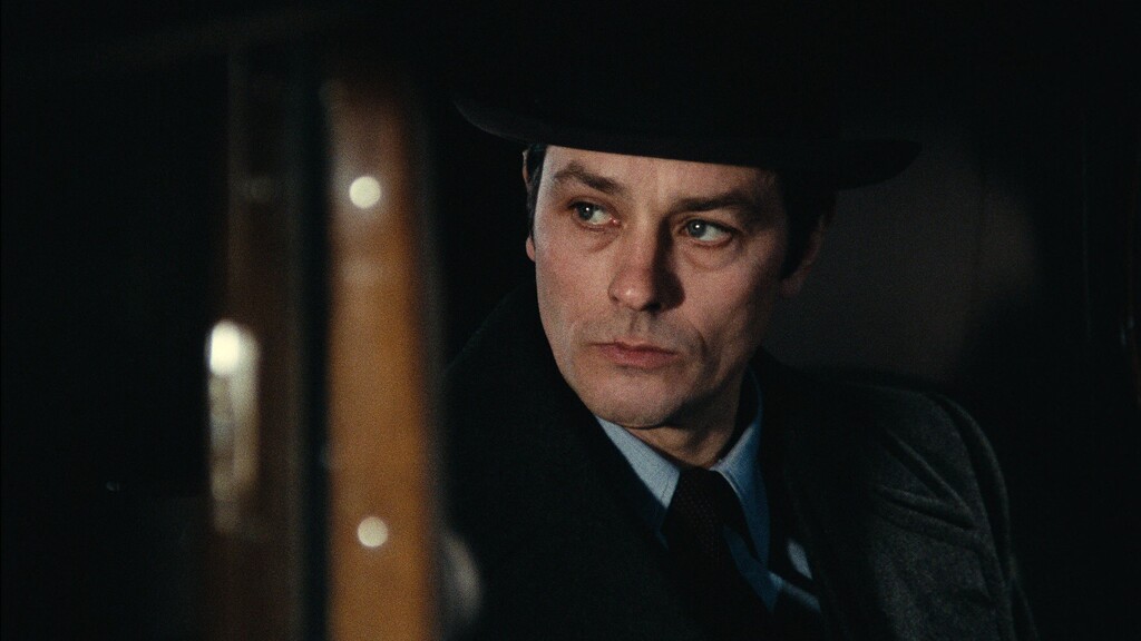 Alain Delon en imperméable et chapeau. L'ambiance évoque un film à suspense.