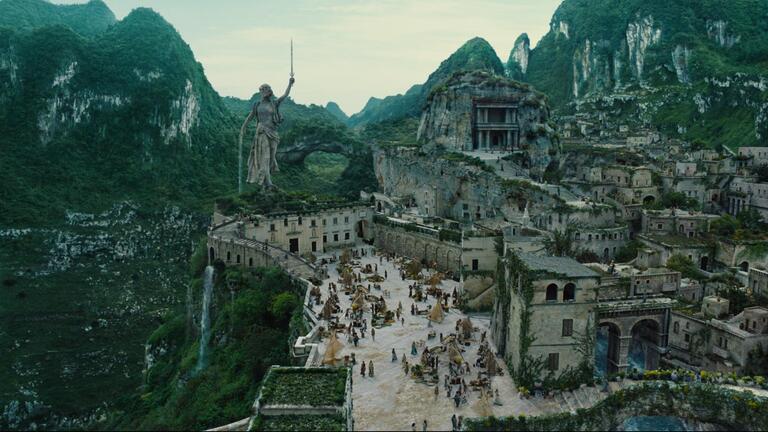 Vue panoramique de Themyscira montrant une cité monumentale intégrée à un relief montagneux, dominée par une statue colossale et organisée autour d’une grande place centrale animée.