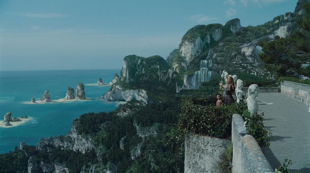 Vue en hauteur du littoral de Themyscira, associant falaises escarpées, végétation dense et formations rocheuses en mer, avec une architecture discrètement intégrée au paysage.