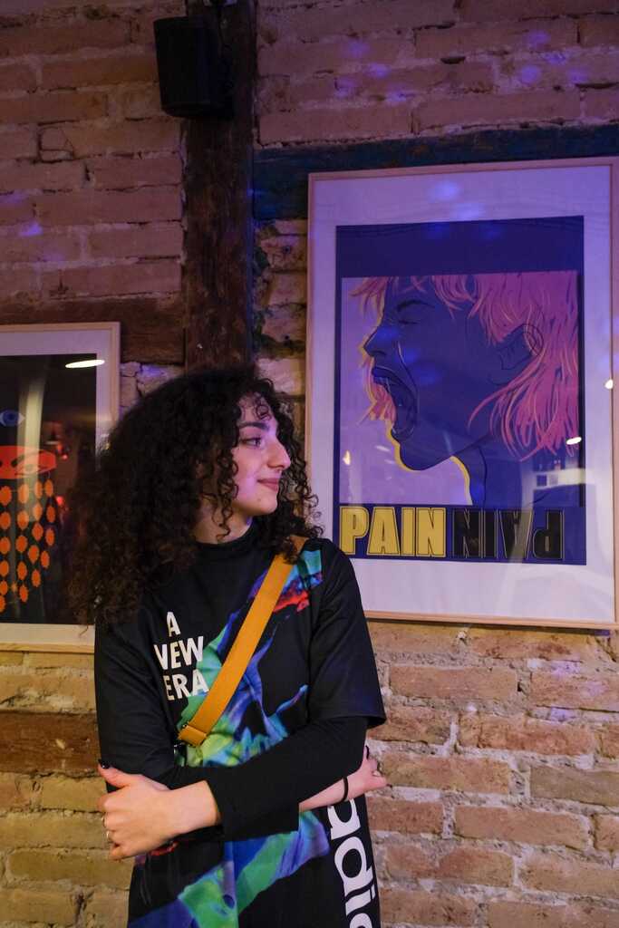 Yona Rouach a gauche de l'image tourne sa tête vers une de ses illustrations exposée.