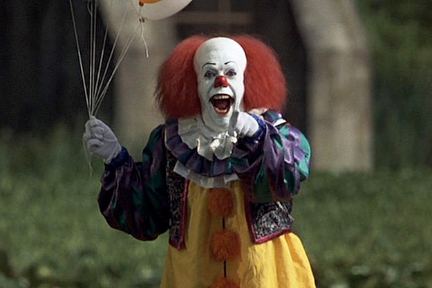 Le clown Pennywise montre du doigt le spectateur, en ouvrant grand la bouche. Il tient des ballons dans sa deuxième main.