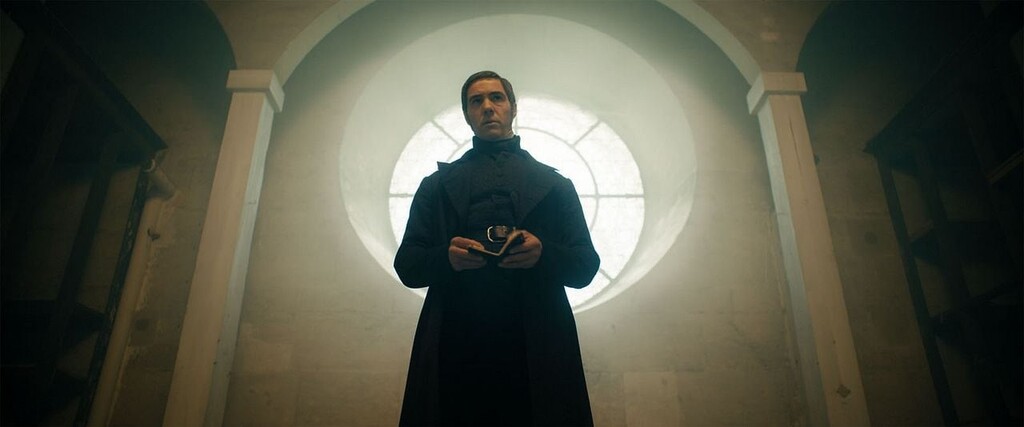 Javert habillé tout en noir. Comme a un carnet dans les mains et se trouve dans un bâtiment austère, il ressemble un peu à un prêtre.