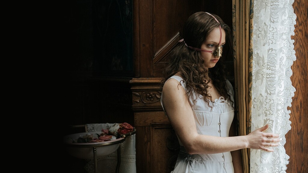Jeune fille en robe blanche et à l'air sombre. Elle a une pièce de métal attachée sur son nez, comme un instrument de chirurgie esthétique barbare. L'ironie est que l'actrice serait très belle sans cette sorte de masque qui la rend effrayante.